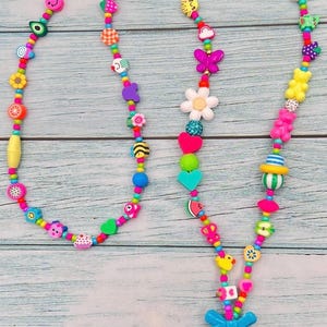 May include: A long lanyard featuring a vibrant assortment of multi-colored plastic beads in various shapes, including flowers, hearts, butterflies, fruits, and smiley faces. The strand ends with a bright blue bow, a pink bead, and a gold metal ring.