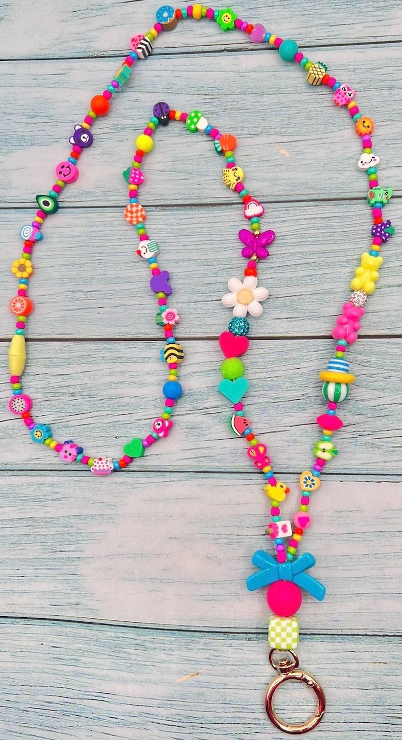 May include: A long lanyard featuring a vibrant assortment of multi-colored plastic beads in various shapes, including flowers, hearts, butterflies, fruits, and smiley faces. The strand ends with a bright blue bow, a pink bead, and a gold metal ring.