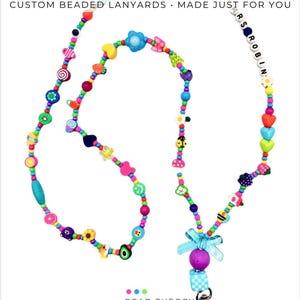 May include: A colorful beaded lanyard featuring a variety of shapes including hearts, flowers, fruits, and a ladybug. The beads are strung on a cord and include a section with the text MRS ROBIN. A light blue bow and a metal clasp are at the bottom.