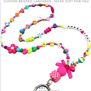 May include: A custom beaded lanyard featuring a variety of colorful shapes including flowers, hearts, smiley faces, and fruits. The text "AMBER CPH" is spelled out in white letter beads. A pink bow and a silver metal lobster clasp are attached.