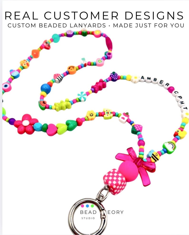 May include: A custom beaded lanyard featuring a variety of colorful shapes including flowers, hearts, smiley faces, and fruits. The text "AMBER CPH" is spelled out in white letter beads. A pink bow and a silver metal lobster clasp are attached.