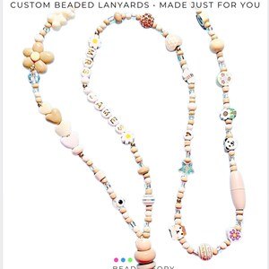 May include: A custom beaded lanyard featuring a variety of light-colored wooden, plastic, and metallic beads in shapes including flowers, hearts, stars, and a panda face. The beads spell out MRS JAMES and include a metal lobster clasp at the bottom.