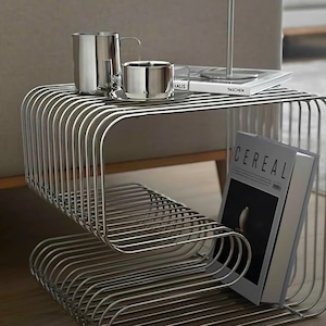May include: A modern, silver-toned metal side table with a unique, curved wireframe design. The table holds two stainless steel mugs, a small pitcher, and a book with the word "HAUS" visible. A book with the word "TASCHEN" is also visible.