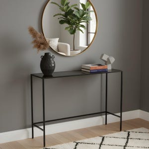 May include: A black metal console table with a rectangular glass top. A black vase with dried flowers, two books, and a white abstract sculpture are on the table. A round gold mirror hangs on the grey wall above the table.