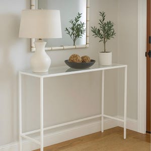 Könnte beinhalten: Ein weißer Metallkonsolentisch mit Glasplatte, auf dem eine weiße Lampe, eine schwarze Schale mit dekorativen Kugeln und eine Topfpflanze stehen. Über dem Tisch hängt ein Spiegel mit Bambusrahmen. Der Raum hat neutrale Wände und einen Holzboden.