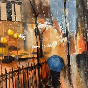 May include: Oil painting depicting a rainy street scene. A person with a blue umbrella walks along a wrought iron fence. Buildings and streetlights are visible in the background, with warm orange and yellow tones contrasting with the cool blues and greys of the rain.