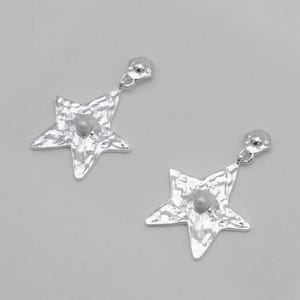 Pu&ograve; includere: Un paio di orecchini a forma di stella color argento. Ogni orecchino presenta una superficie strutturata e una piccola perla rotonda di colore chiaro al centro. Gli orecchini pendono da un piccolo perno rotondo.