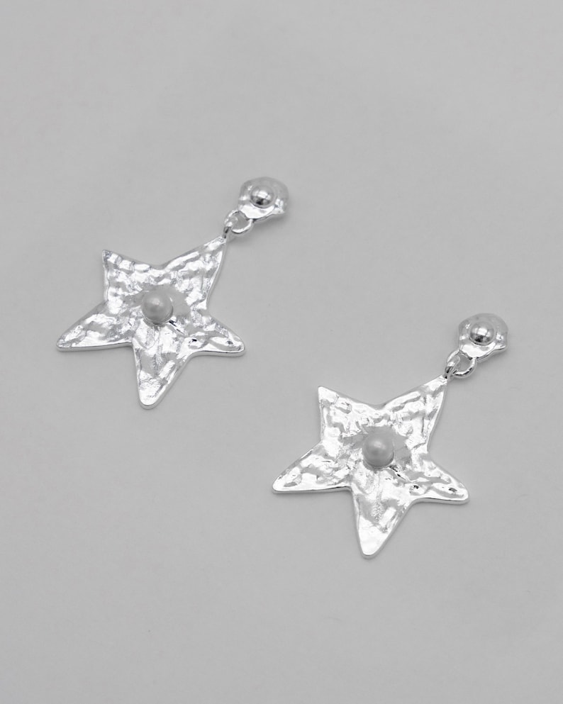 Pu&ograve; includere: Un paio di orecchini a forma di stella color argento. Ogni orecchino presenta una superficie strutturata e una piccola perla rotonda di colore chiaro al centro. Gli orecchini pendono da un piccolo perno rotondo.