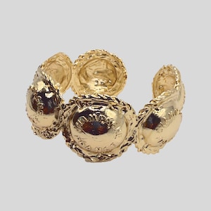 Könnte beinhalten: Ein goldfarbenes Armband, das aus mehreren miteinander verbundenen kreisförmigen Medaillons besteht. Jedes Medaillon hat einen strukturierten, seilartigen Rand und den geprägten Schriftzug "CHAMPAGNE FRANCE" in der Mitte, was ein durchgehendes Metalldesign ergibt.