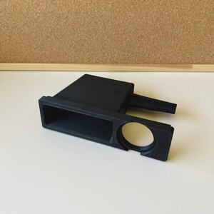 Puede incluir: Un accesorio negro mate impreso en 3D que cuenta con un compartimento de almacenamiento rectangular y una abertura circular en el lateral. El objeto tiene una base plana y un brazo que sobresale hacia atrás, con un acabado texturizado.