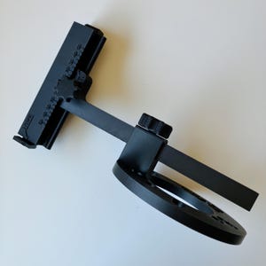 May include: A black 3D-printed cup turner attachment featuring a circular base with mounting holes, a central adjustable arm, and a horizontal bracket with a printed scale labeled SIZE, 10, 20, 30, and 40, secured by two black plastic star knobs.
