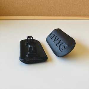 May include: Two matte black plastic automotive interior components. One piece shows the top surface with the embossed text CIVIC, while the other piece is flipped over to reveal the integrated plastic mounting clips on the underside.