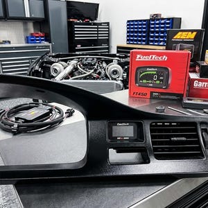 May include: A black automotive dashboard gauge pod insert featuring a FuelTech digital display screen and a horizontal air vent. The unit includes a black wiring harness with connectors and is positioned on a workbench near a FuelTech FT450 box.