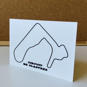 Op de afbeelding: Een wit rechthoekig bord met een zwarte draadomtrek van het racecircuit Circuit de Clastres. De tekst "CIRCUIT DE CLASTRES" is in zwarte, vette, schreefloze hoofdletters onder het circuitontwerp op het witte oppervlak gedrukt.