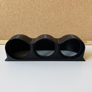 Op de afbeelding: Een matzwarte triple meterhouder met drie ronde openingen in een horizontale rij. De structuur heeft een getextureerde afwerking en bevat een platte basis voor montage, ontworpen om drie ronde meters in te plaatsen.