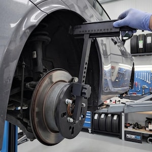 Op de afbeelding: Een zwart metalen spatbordrolgereedschap bevestigd aan een wielnaaf van een auto. Het gereedschap is voorzien van een horizontale meetschaal met markeringen van 0 tot 50 en een verticale arm met een knop voor het meten van de ruimte in de wielkast.