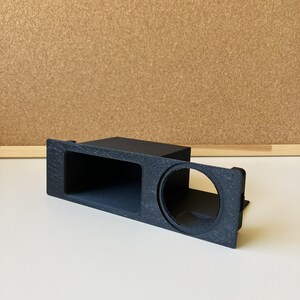 May include: A matte black rectangular console insert featuring a wide, open storage compartment on the left side and a circular cup holder opening on the right side, constructed with a textured finish and rigid, angular edges.