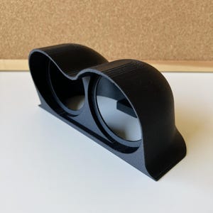Può includere: Un supporto doppio per manometri stampato in 3D in nero opaco con due aperture circolari per la strumentazione automobilistica. La struttura presenta una superficie superiore curva e sagomata con linee di strato visibili e una base piatta.