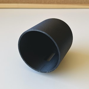 Op de afbeelding: Een matzwart cilindervormig object met een holle binnenkant en een getextureerd oppervlak. Het item heeft een enkele rechthoekige uitsparing nabij de basis van de zijwand, met een consistente donkere afwerking over de gehele buitenstructuur.