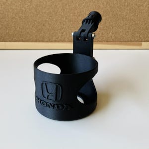 May include: A matte black plastic cup holder featuring a cylindrical body with a circular cutout and the Honda logo embossed on the front. The holder includes an adjustable mounting arm with a hinge mechanism and a small silver metal bolt.