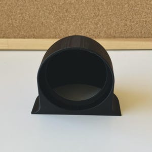 Op de afbeelding: Een matzwarte, 3D-geprinte ronde meterhouder met een hol cilindrisch midden en een platte, rechthoekige basis. Het onderdeel heeft een gladde, uniforme afwerking en is ontworpen om een rond instrument of meter stevig op zijn plek te houden.