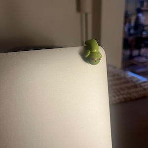 Op de afbeelding: Een kleine, groene kikkerfiguur met rode accenten, geplaatst op de hoek van een zilveren laptop. De kikker heeft een afgerond lichaam en een glimlachende uitdrukking. Het figuur is gemaakt van een glad materiaal.