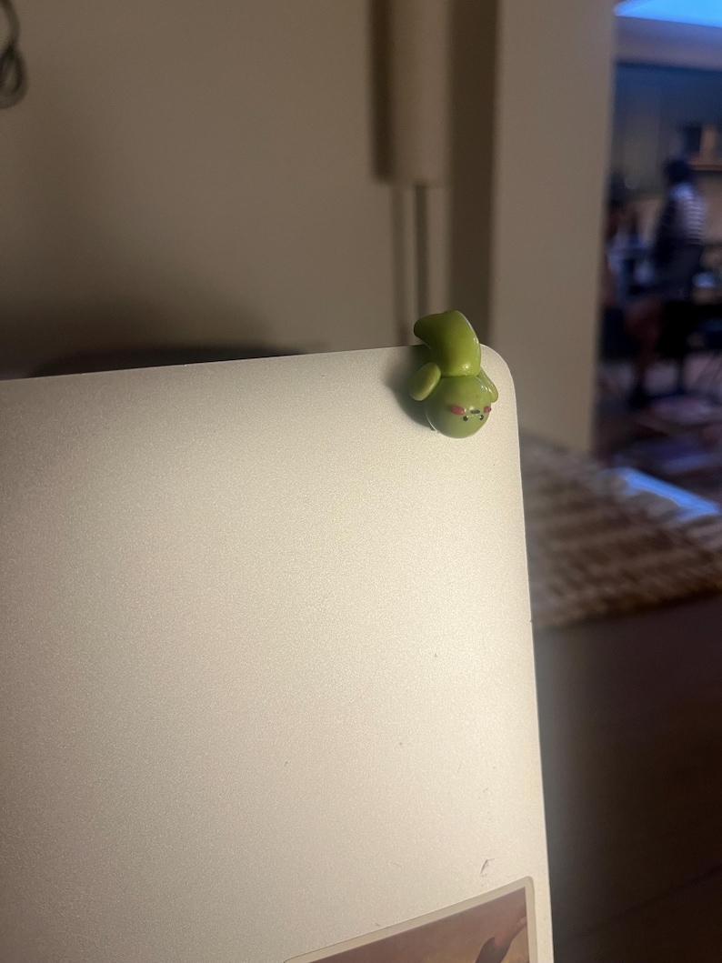 Op de afbeelding: Een kleine, groene kikkerfiguur met rode accenten, geplaatst op de hoek van een zilveren laptop. De kikker heeft een afgerond lichaam en een glimlachende uitdrukking. Het figuur is gemaakt van een glad materiaal.