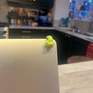 Op de afbeelding: Een klein, limoengroen, cartoonachtig wezen is bevestigd aan de hoek van een zilveren laptop. Het wezen heeft een afgerond lichaam, kleine ogen en een lachende mond. De achtergrond is een keuken.