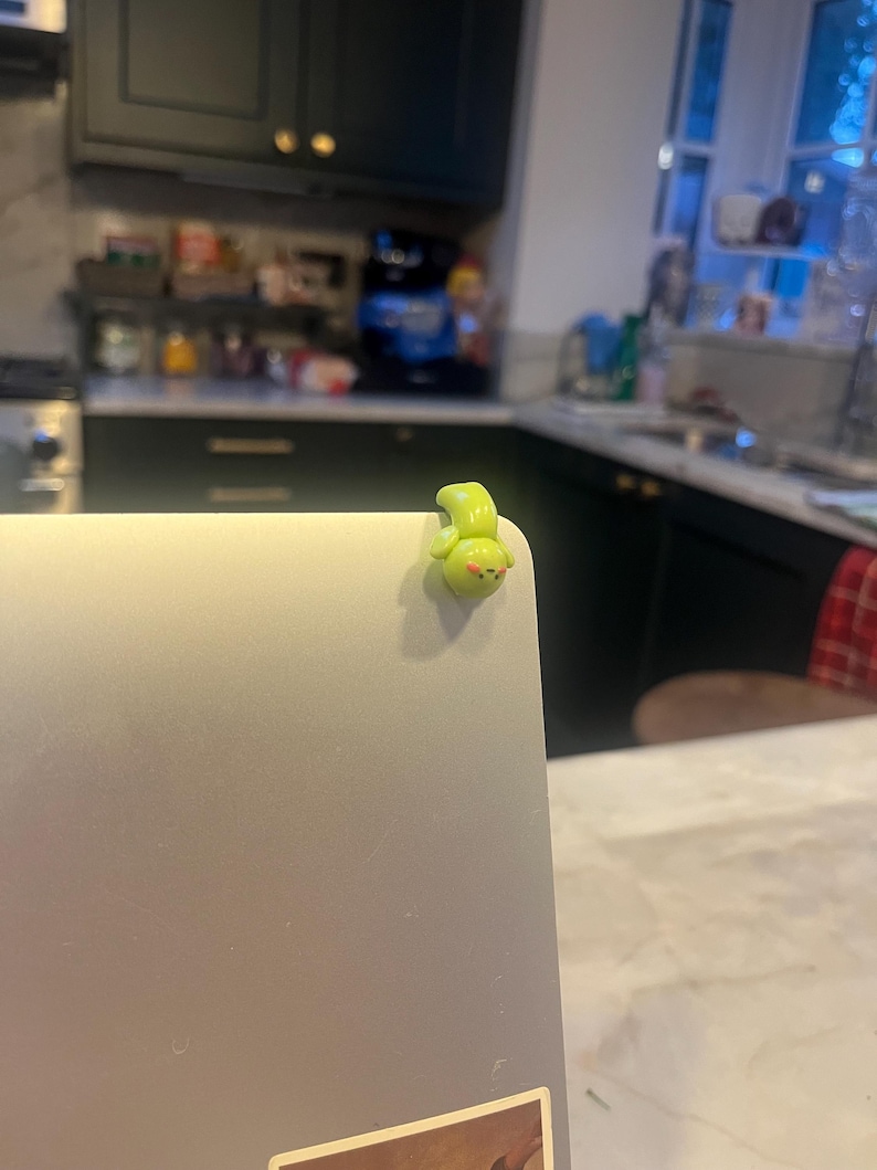 Op de afbeelding: Een klein, limoengroen, cartoonachtig wezen is bevestigd aan de hoek van een zilveren laptop. Het wezen heeft een afgerond lichaam, kleine ogen en een lachende mond. De achtergrond is een keuken.