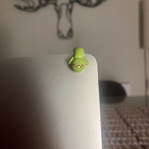 Op de afbeelding: Een kleine, lichtgroene, handgemaakte kleifiguur van een stripfiguur met een lachend gezicht en kleine armen. Het beeldje is bevestigd aan de hoek van een zilveren laptop. De achtergrond is wazig, met een tekening van geweien aan de muur.
