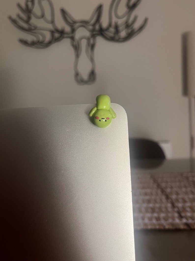 Op de afbeelding: Een kleine, lichtgroene, handgemaakte kleifiguur van een stripfiguur met een lachend gezicht en kleine armen. Het beeldje is bevestigd aan de hoek van een zilveren laptop. De achtergrond is wazig, met een tekening van geweien aan de muur.
