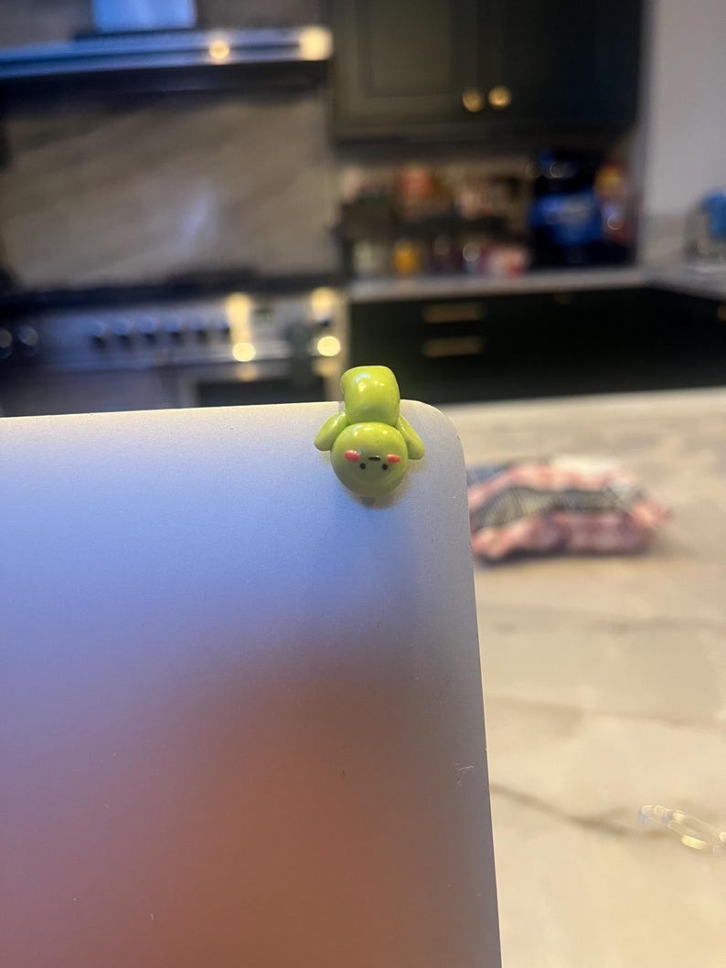 Op de afbeelding: Een kleine, groene figuur in cartoonstijl met een rond gezicht en kleine armen is bevestigd aan de hoek van een laptop. De figuur heeft een lichtgroen lichaam, een roze gezicht en zwarte ogen. De laptop is zilver.