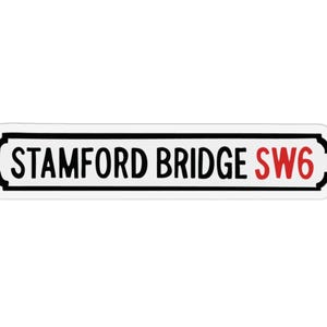 May include: A white sticker with a black border and the words "STAMFORD BRIDGE SW6" printed on it. The text "STAMFORD BRIDGE" is in black, and "SW6" is in red. The sticker has a rectangular shape with rounded edges.