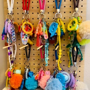 May include: A collection of colorful keychains with various charms, including pom-poms, beads, and star shapes. Each keychain has a different colored lanyard. The keychains are displayed on a wooden pegboard with black hooks.