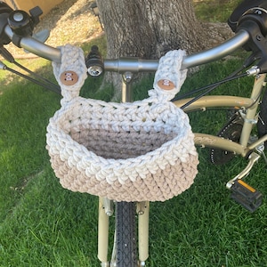 Könnte beinhalten: Ein beige-weißer gehäkelter Korb ist am Lenker eines Fahrrads befestigt. Der Korb ist aus dickem Garn gefertigt und hat ein schlichtes, modernes Design.