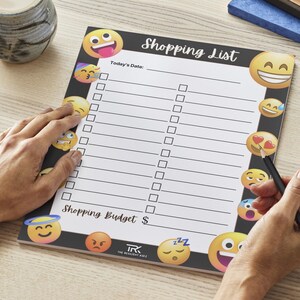May include: A black and white shopping list notepad with the words "Shopping List" at the top. The notepad has a series of emoji faces around the border. A hand is holding a pencil and writing on the notepad.