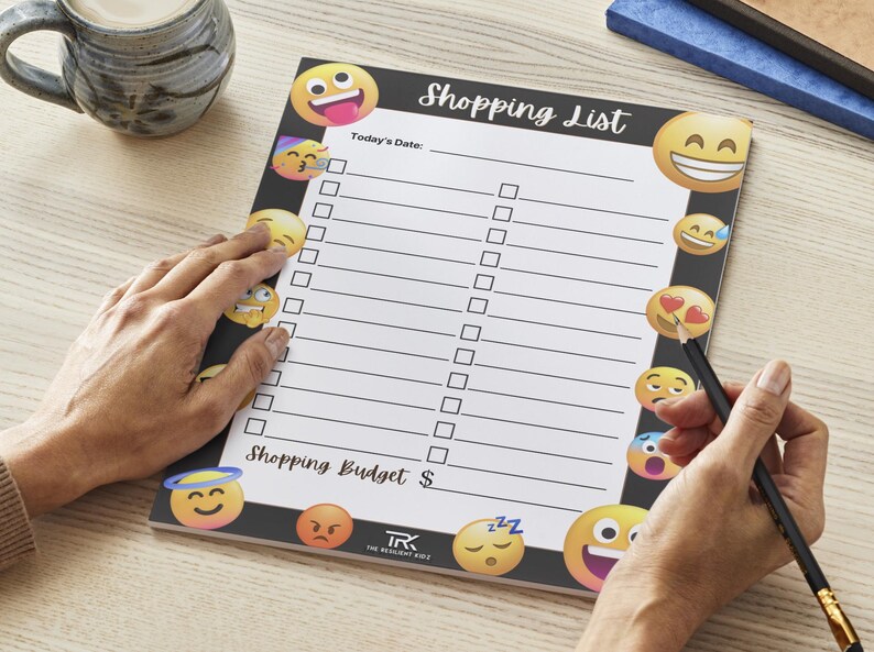 May include: A black and white shopping list notepad with the words "Shopping List" at the top. The notepad has a series of emoji faces around the border. A hand is holding a pencil and writing on the notepad.