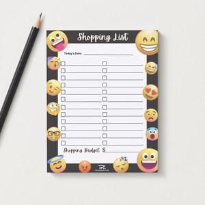 May include: A black shopping list notepad with a border of emoji faces. The notepad has a header that says "Shopping List" and includes spaces for the date and budget. A black pencil lies to the left of the notepad.