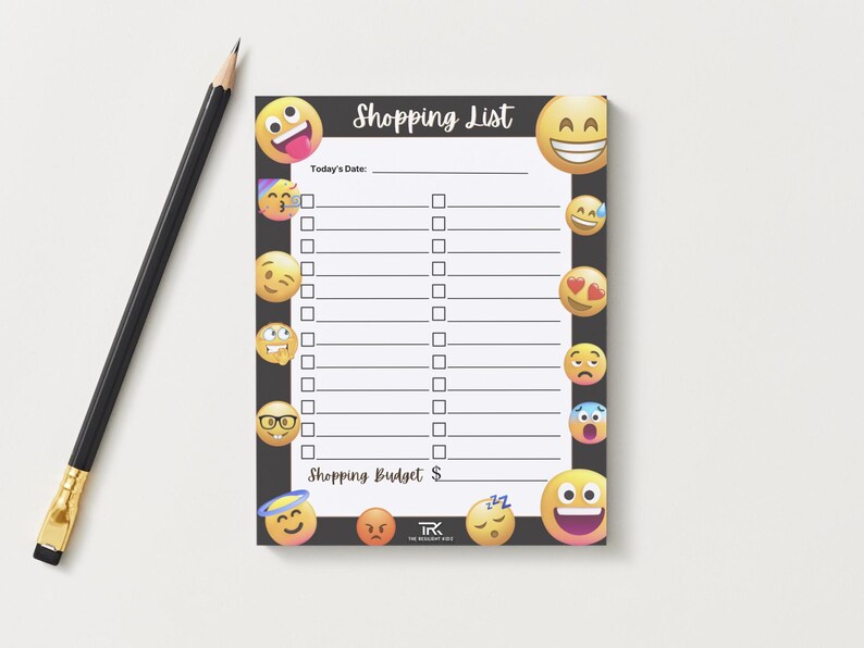 May include: A black shopping list notepad with a border of emoji faces. The notepad has a header that says "Shopping List" and includes spaces for the date and budget. A black pencil lies to the left of the notepad.