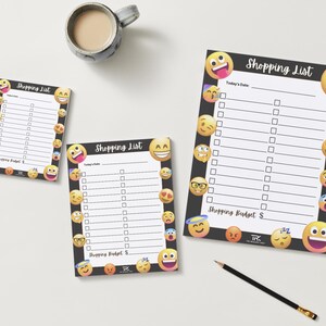 May include: Three shopping list notepads with a black border and emoji designs. The notepads have a white interior with lines and check boxes. The text "Shopping List" is at the top of each notepad. A pencil and a mug of coffee are also in the image.