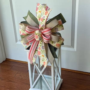 May include: A white lantern topped with a decorative bow. The bow incorporates ribbons with floral, polka dot, and striped patterns. The colour scheme includes pink, green, beige, and white. The lantern is constructed from wood and has a glass enclosure.