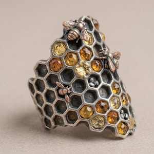 May include: A silver ring shaped like a honeycomb with a dark oxidised finish. The ring features several small, faceted yellow and amber-coloured gemstones set within the hexagonal cells, along with three small, detailed copper-coloured bee figures.