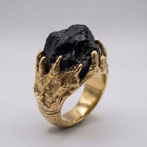 Pode incluir: Anel dourado com uma grande pedra preta bruta. A cravação do anel apresenta um design texturizado e orgânico, que se assemelha a garras. A pedra preciosa é de cor escura e opaca, contrastando com o metal dourado.