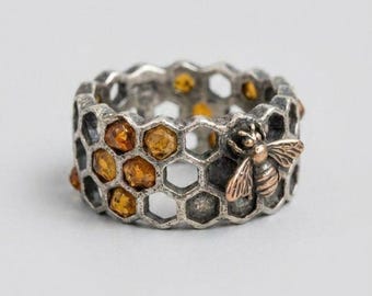 May include: A silver-toned ring featuring a textured honeycomb pattern throughout the band. Several hexagonal cells are filled with small, faceted amber-colored stones, and a small metallic bee figure is attached to the side of the band.