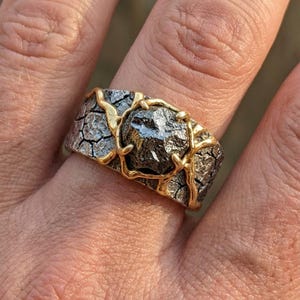 Könnte beinhalten: Ein einzigartiger Ring mit einem dunklen, unregelmäßig geformten Stein, der von goldfarbenen Metallakzenten gehalten wird. Das Band des Rings hat ein strukturiertes, rissiges Aussehen, wobei das Goldmetall den Stein umschließt.