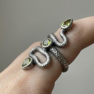 Könnte beinhalten: Ein silberfarbener Ring in Form einer gewundenen Schlange mit gehämmertem Finish. Die Schlange ist mit drei grünen Edelsteinen besetzt: einem runden Stein in der Mitte und zwei tropfenförmigen Steinen an Kopf und Schwanzende.