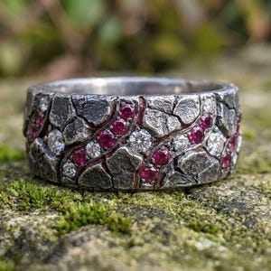 Pode incluir: Um anel largo de prata texturizado com um design de mosaico. O anel é incrustado com pequenas pedras de forma irregular, intercaladas com pedras preciosas vermelhas e claras. O anel está sobre uma superfície musgosa.