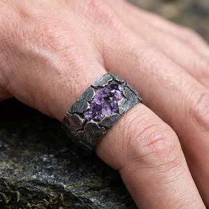 May include: A silver ring with a textured, cracked surface. The centre of the ring is filled with a cluster of small, sparkling purple crystals. The ring is worn on a finger, showcasing the unique design and gemstone.