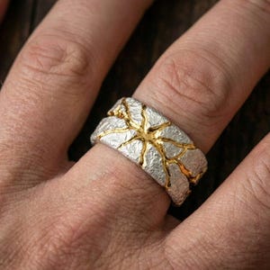 May include: A silver ring with a textured surface and gold-colored accents. The ring features a unique design with gold-colored lines running across the silver band, creating a striking contrast. The ring is shown on a finger.