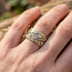 Könnte beinhalten: Ein goldfarbener Ring mit strukturierter Oberfläche und Blattmuster. Der Ring hat kleine, funkelnde Edelsteine, die in die Blattmuster eingesetzt sind. Es ist ein breiter Bandring.